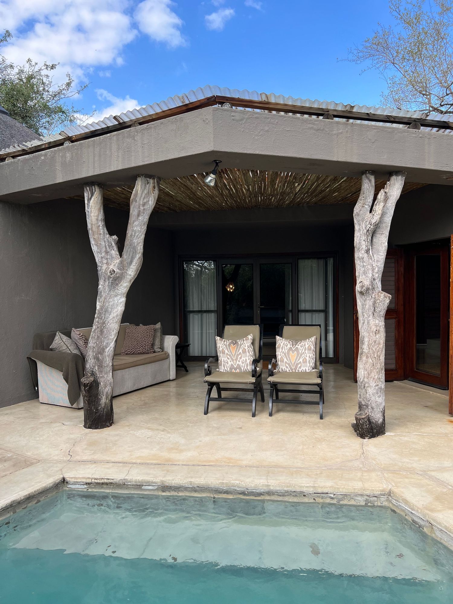 Modern covered patio with rustic tree trunk pillars and poolside lounge seating overlooking a small plunge pool.