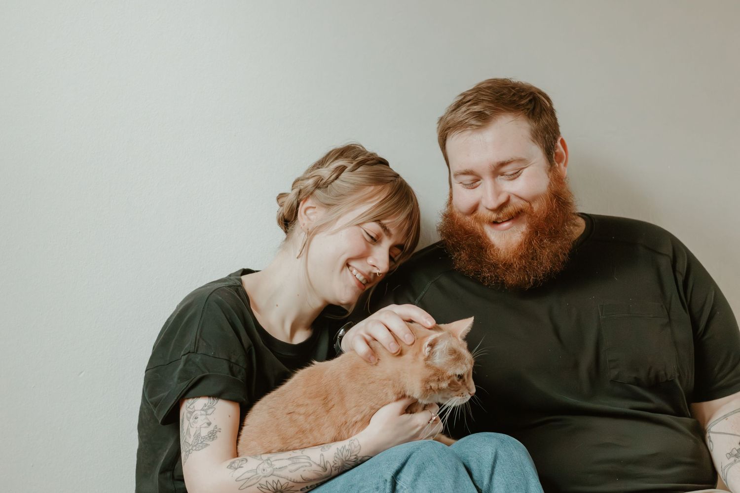 Couple cuddling with their orange cat against a minimalist gray background.