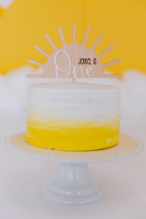 Yellow and white birthday cake on pedestal against bright background.