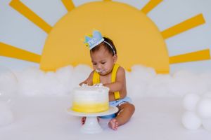 Baby sitting near white cake with yellow sun backdrop during celebration.