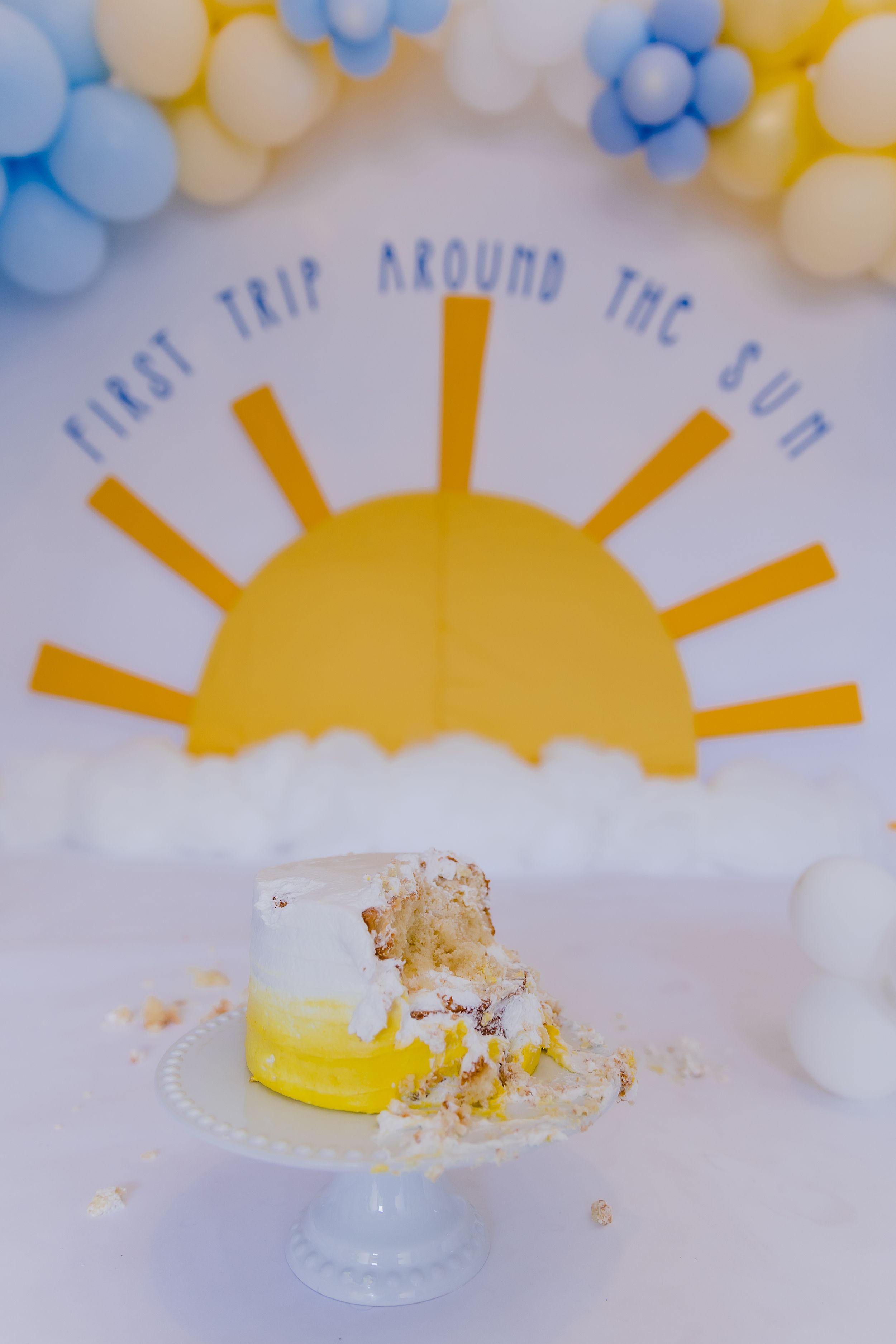 Destroyed yellow cake on white pedestal with balloon decorations.