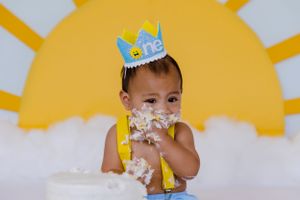 Baby in party hat playing with cake against yellow sun backdrop.