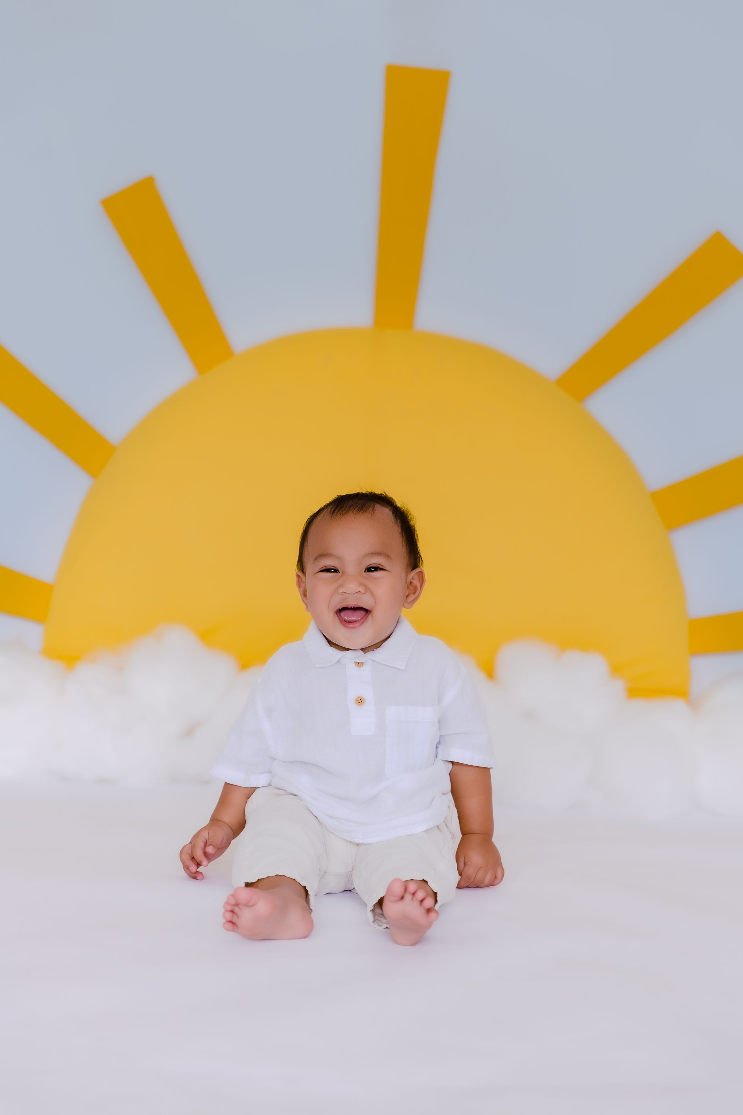 Baby in white outfit lying on floor with yellow sun decoration in background.