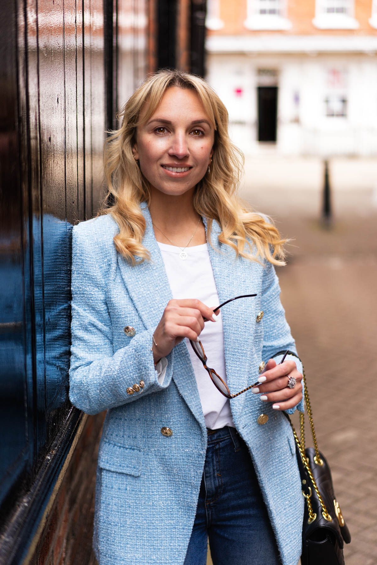 Stylish businesswoman in light blue blazer holding glasses against brick wall.
