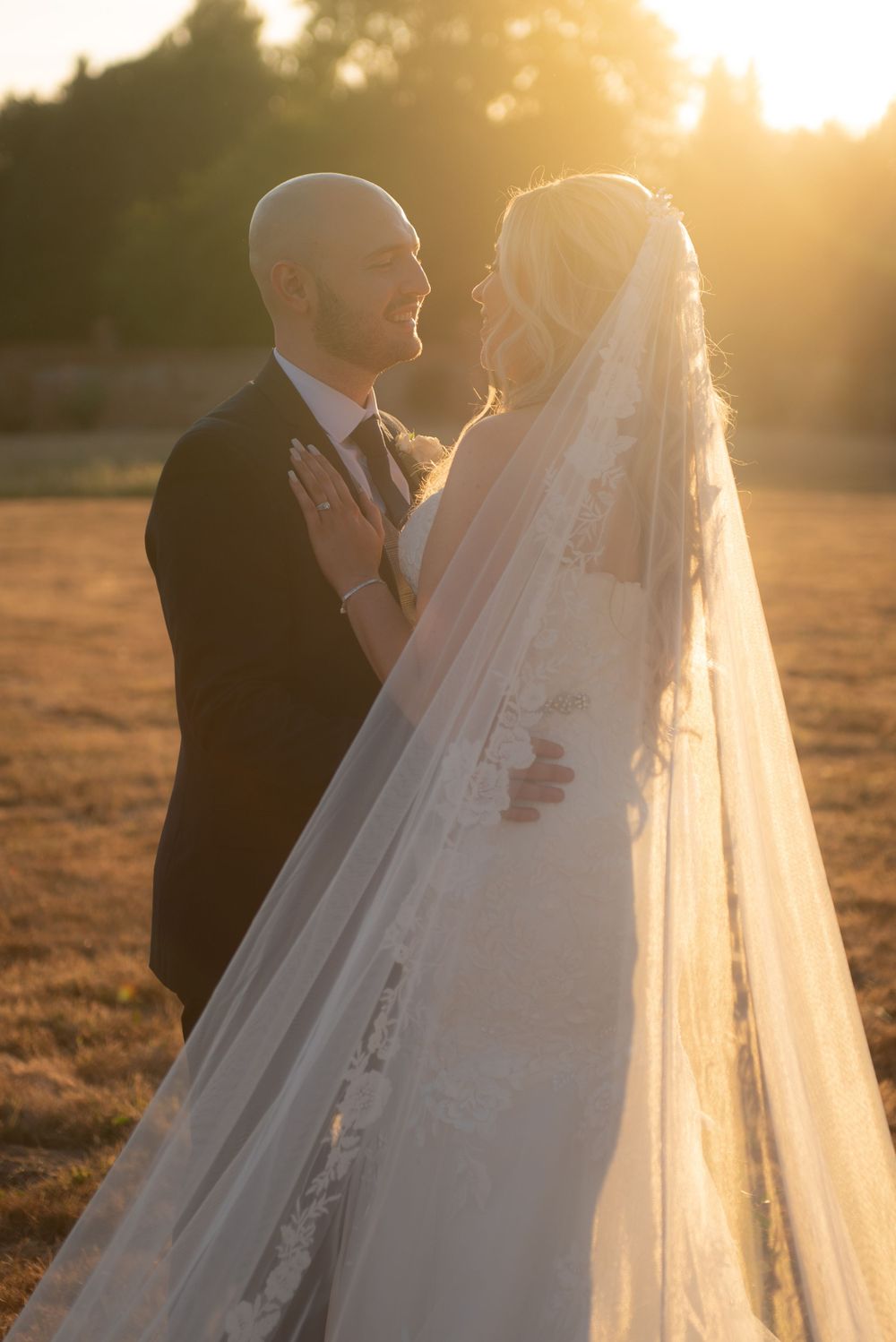 Wedding couple embraces in a golden sunset field with bride wearing long flowing veil.