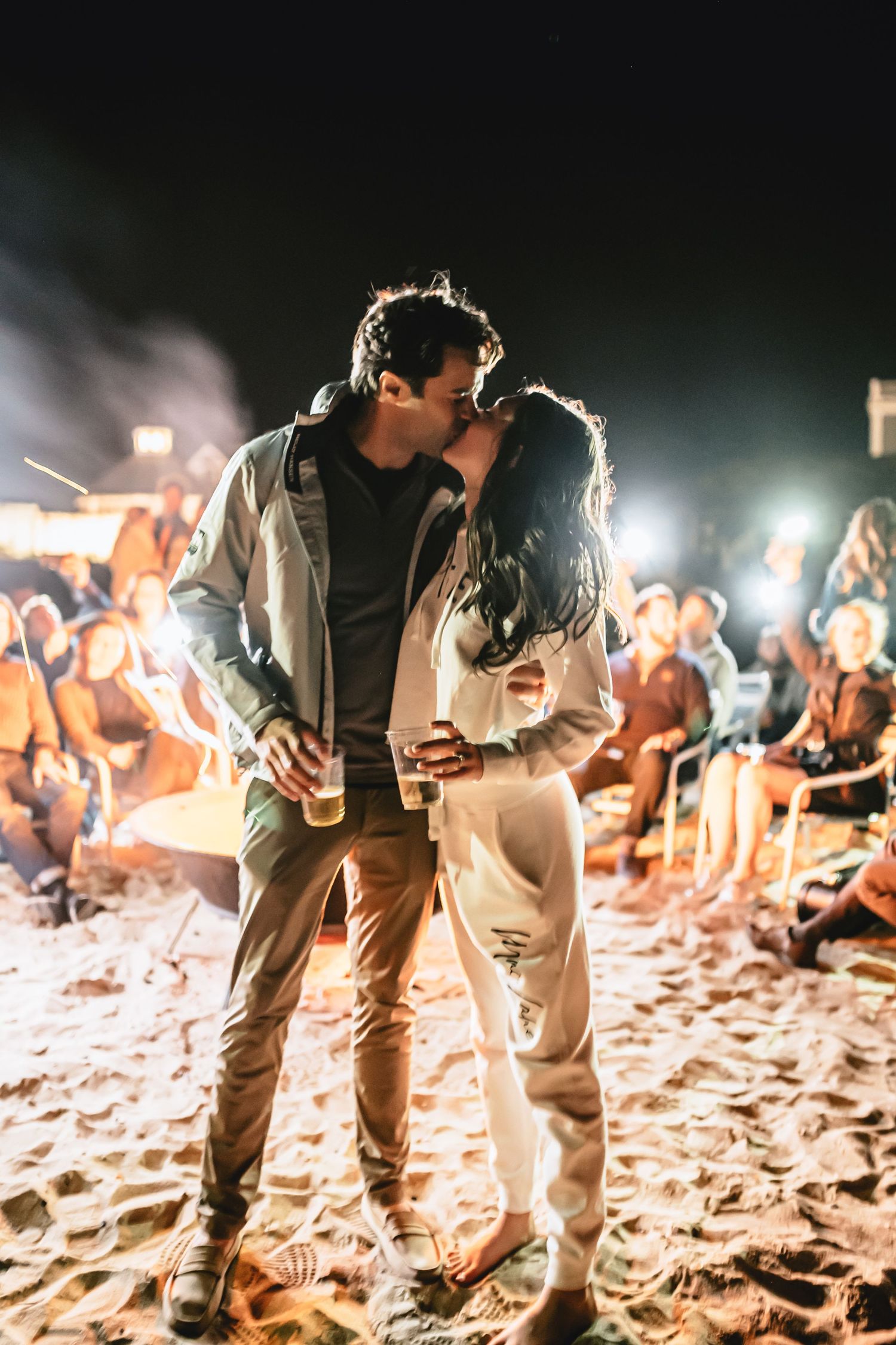 Couple sharing an intimate moment on a beach at night with crowd in the background.