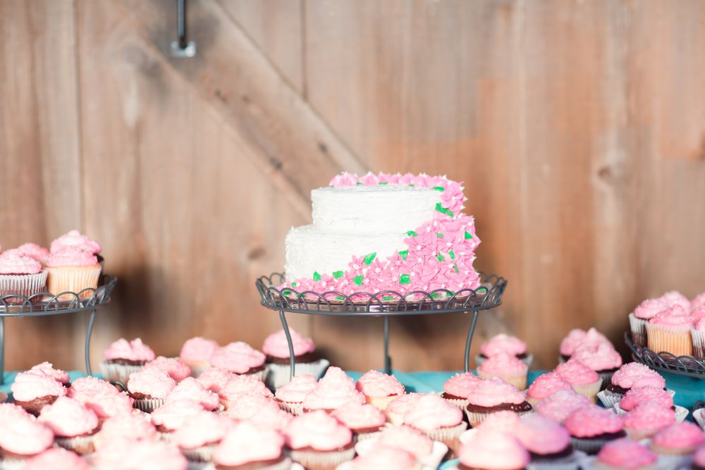 A white wedding cake with pink floral decorations surrounded by pink cupcakes on display.