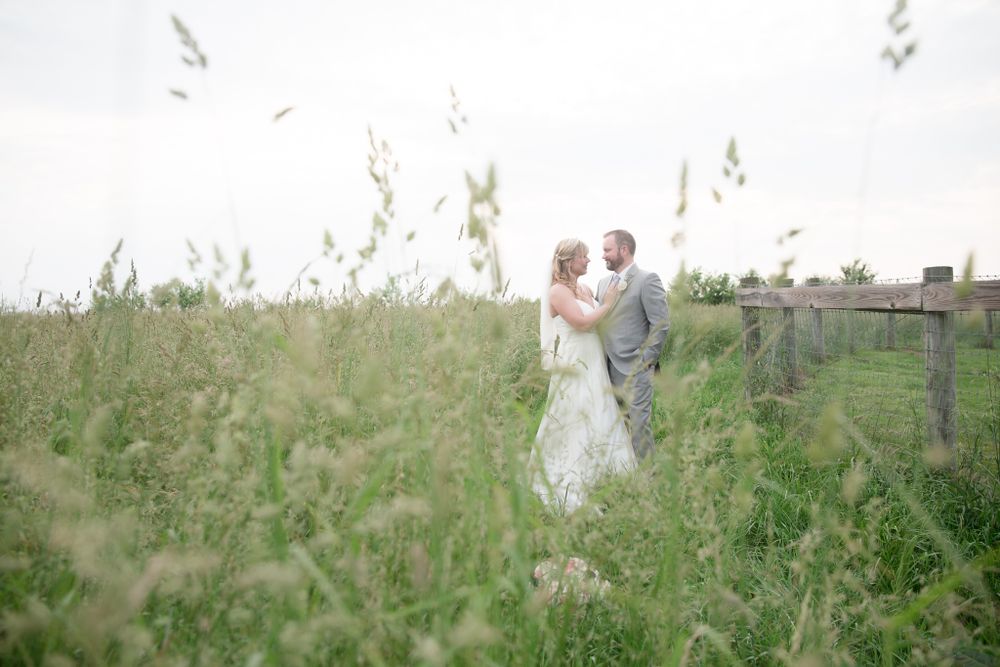 Wedding couple embraces in a dreamy meadow surrounded by tall grasses and rustic fencing.