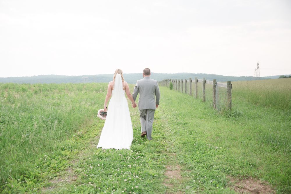 A couple walks hand in hand through a serene grassy field with fence posts along a countryside path.