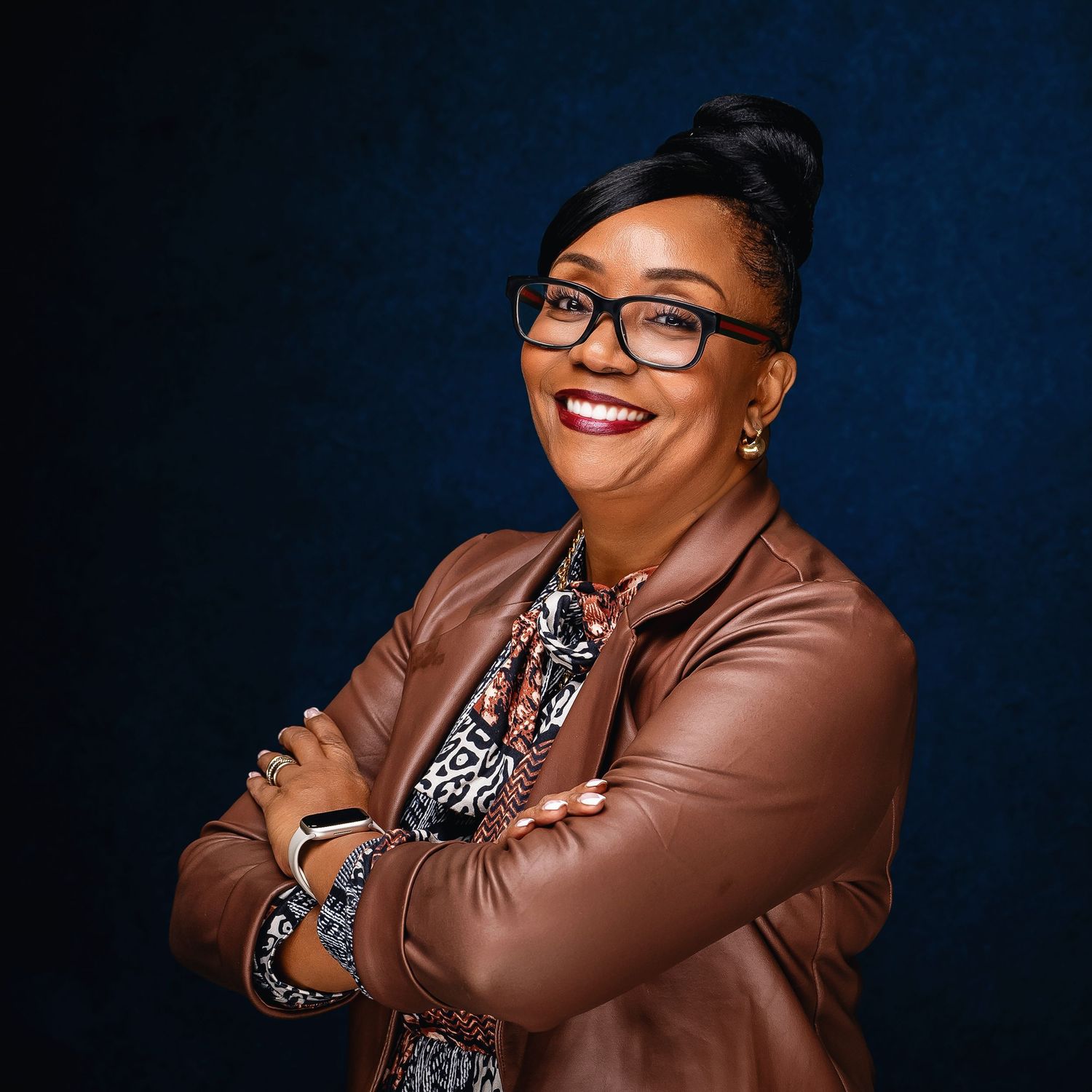 Portrait of person in brown leather jacket and glasses with updo hairstyle against dark background.
