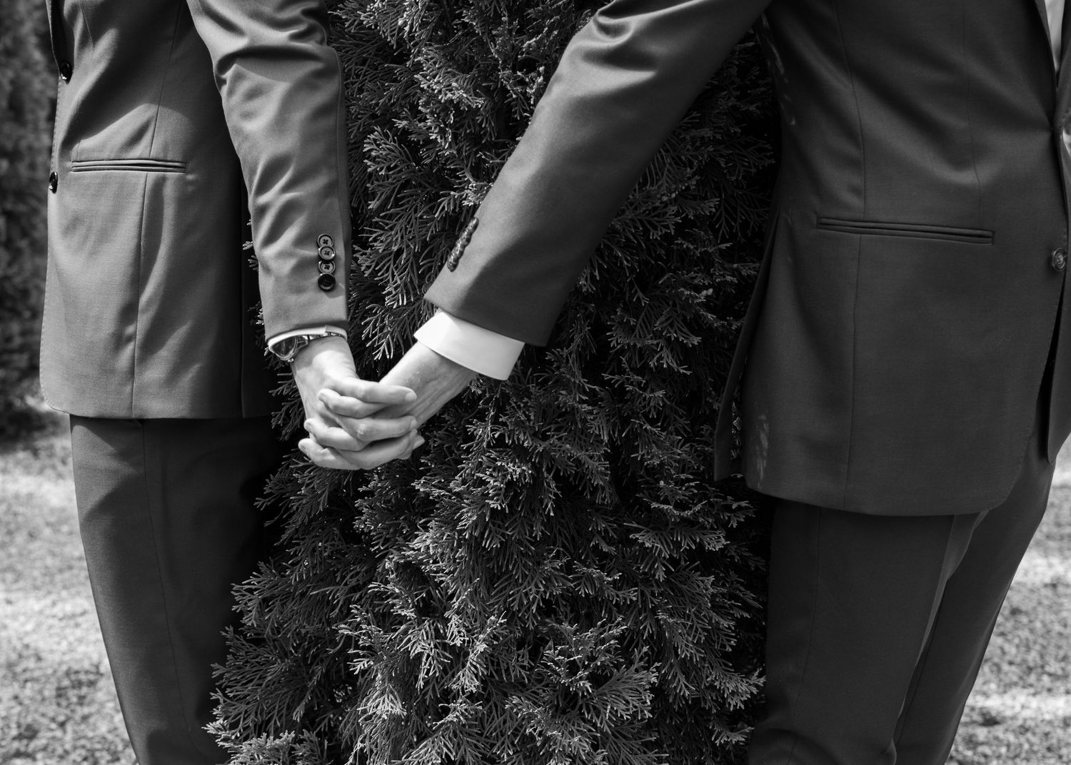 A black and white photo showing hands and legs of two grooms in formal attire on their wedding day, holding handsgathered together outdoors.