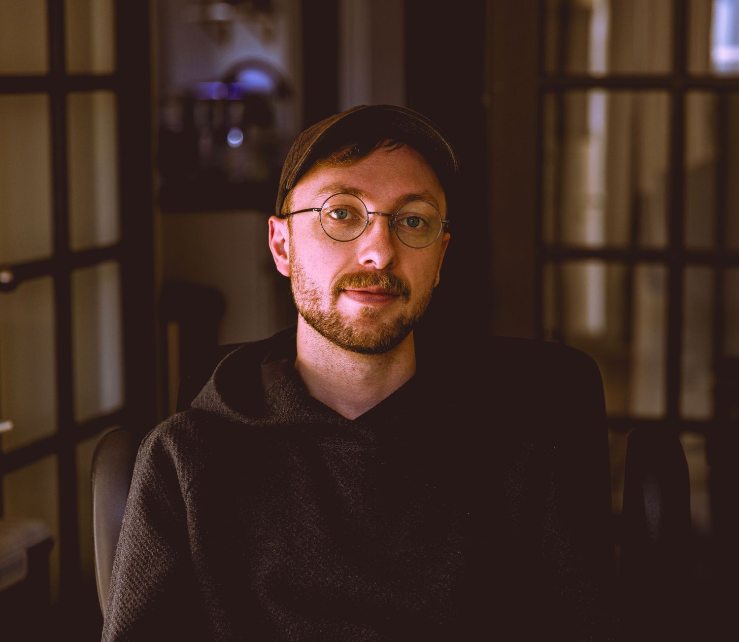 Bearded man with glasses wearing a cap and black sweater, sitting indoors, posed for professional portrait headshots.