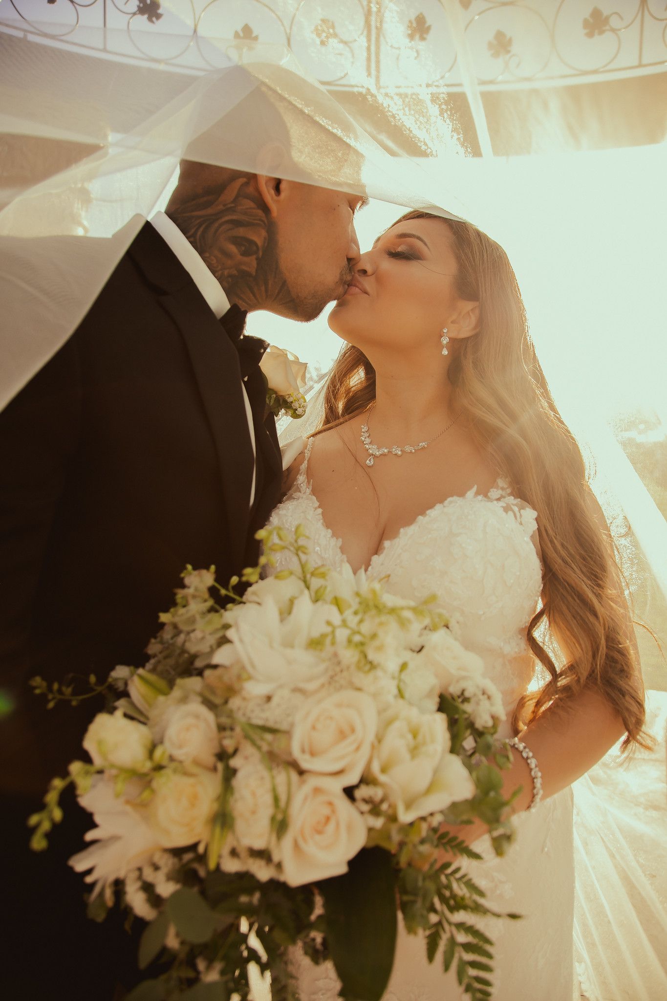 Newlyweds share a kiss under flowing veil with a bouquet of white roses.