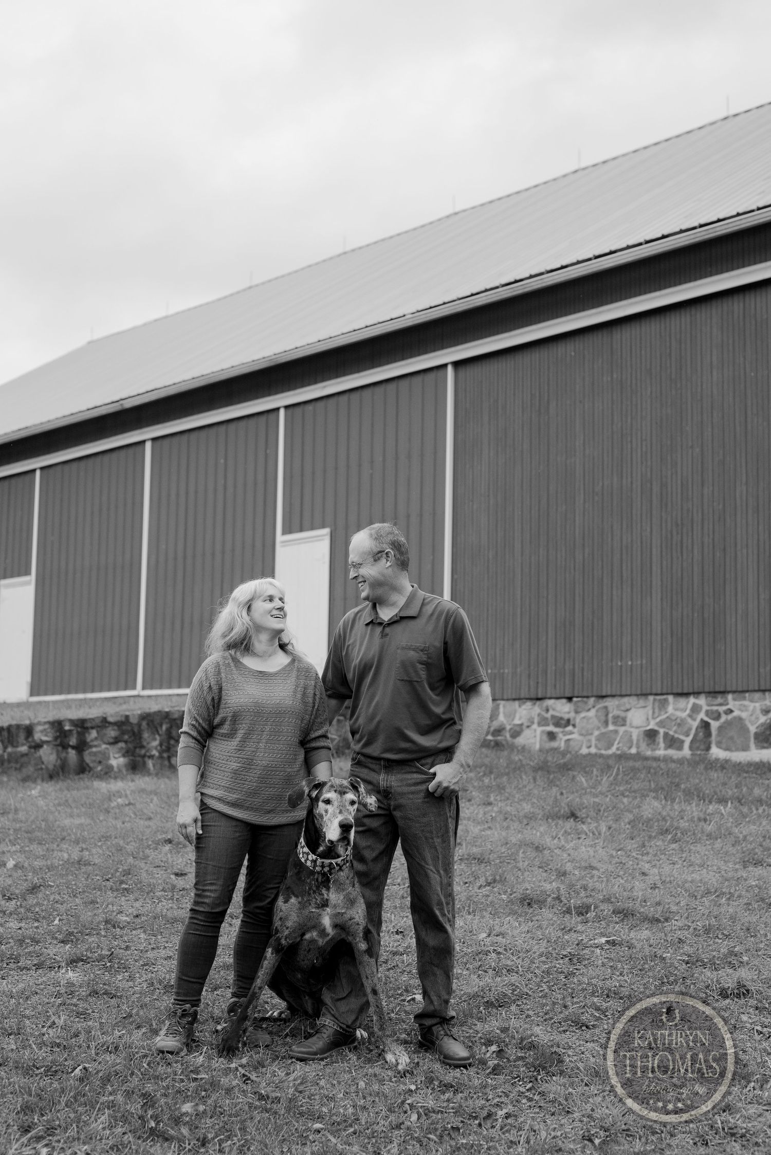 A black and white portrait of a couple with their dog standing by a rustic barn.
