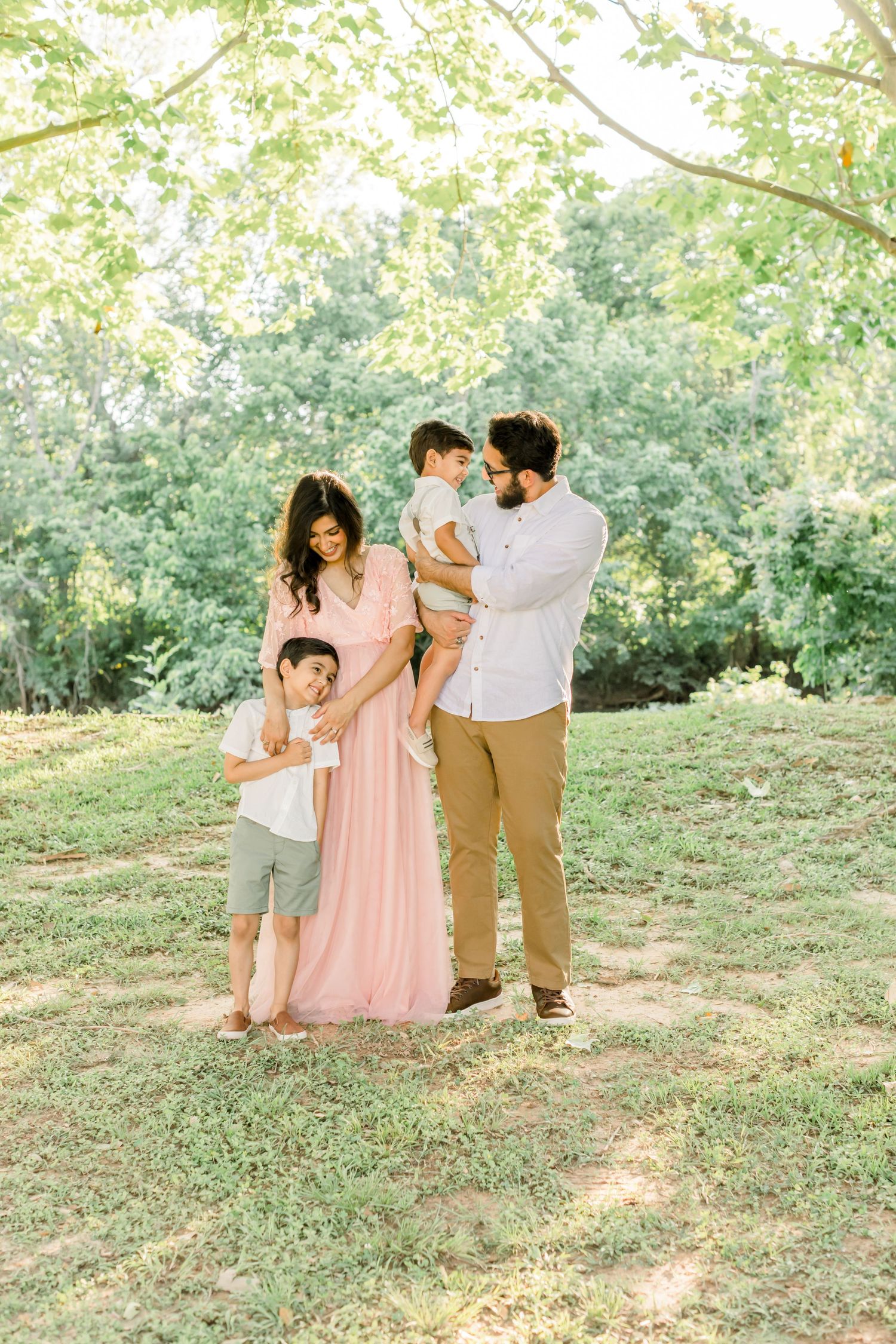 Family poses together on grassy hill wearing coordinated pink and neutral outfits in sunlit outdoor setting.