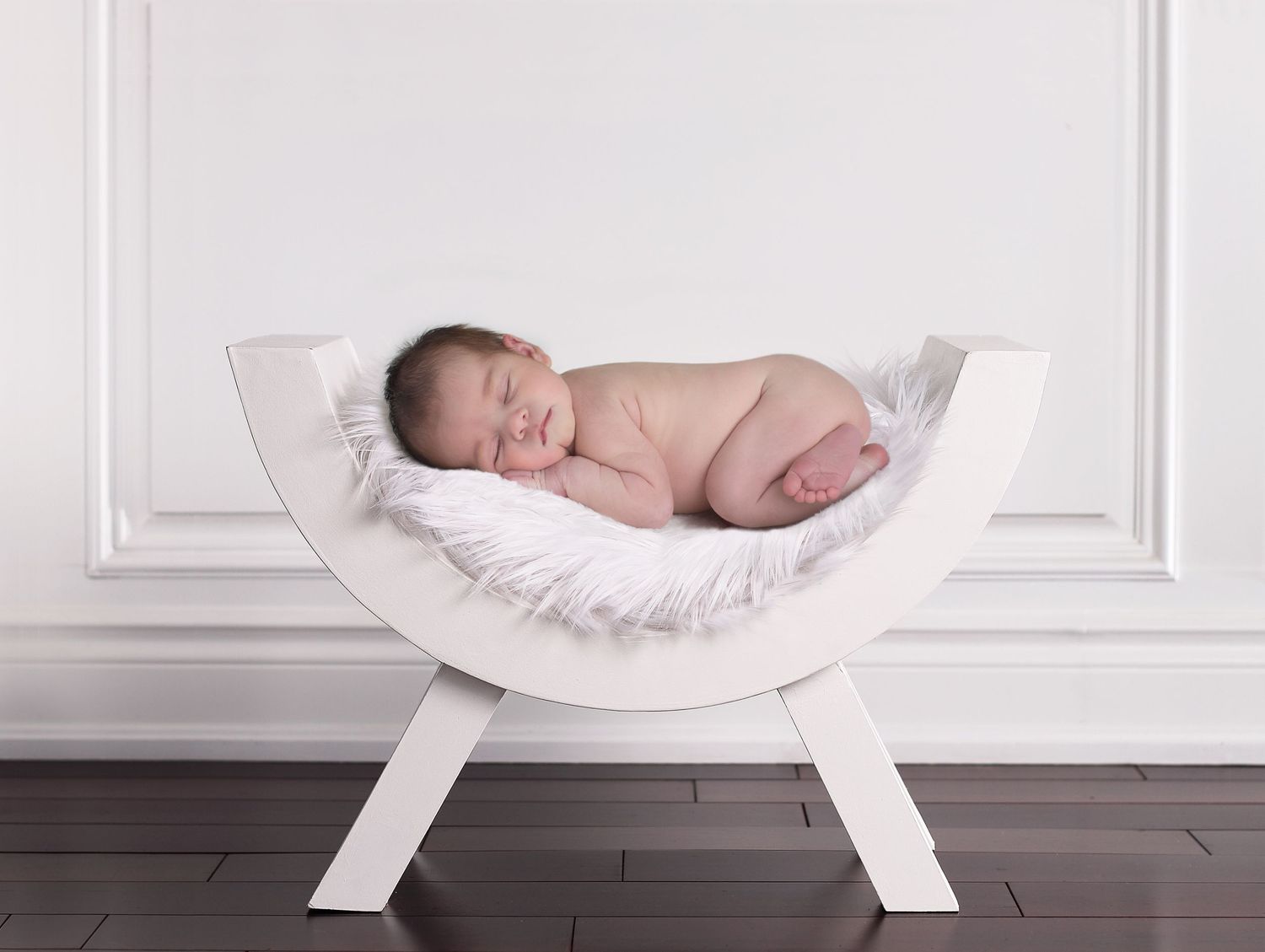 White curved hammock stand displaying sleeping newborn against white paneled wall.