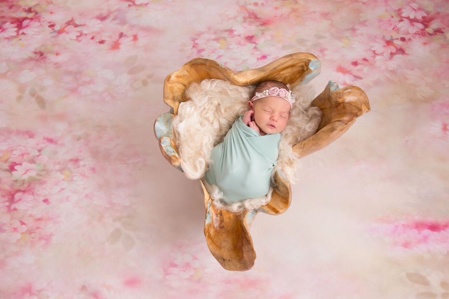 Sleeping infant in mint outfit surrounded by tan blankets on pink floral backdrop.