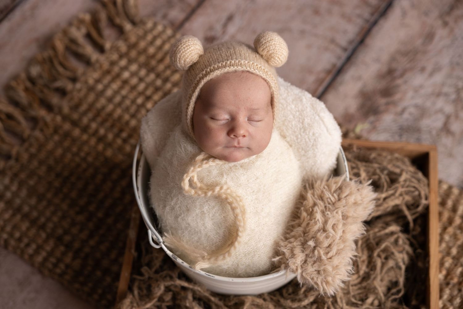 Sleeping baby in cream bear outfit resting in white bowl on textured background.