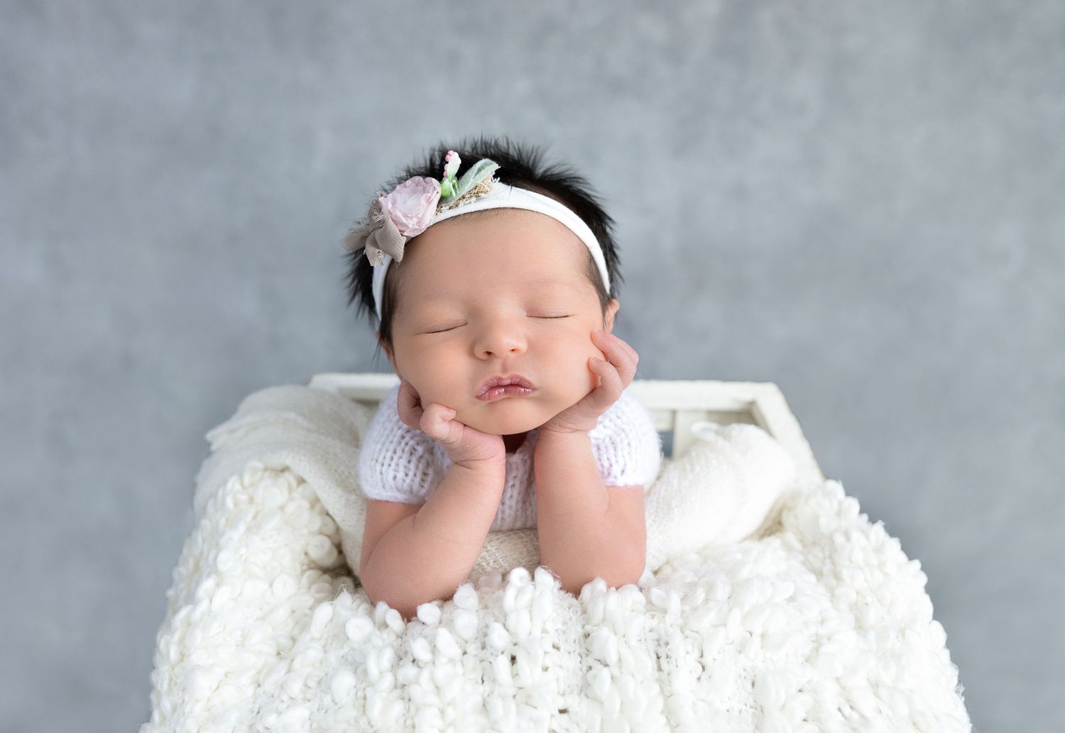 Sleeping baby on white blanket with hands under cheeks and floral headband.
