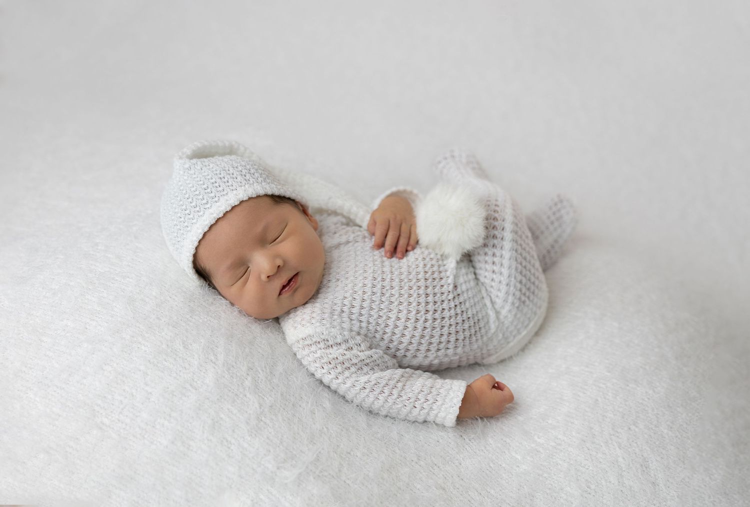 Sleeping newborn wearing white knit outfit on soft white background.