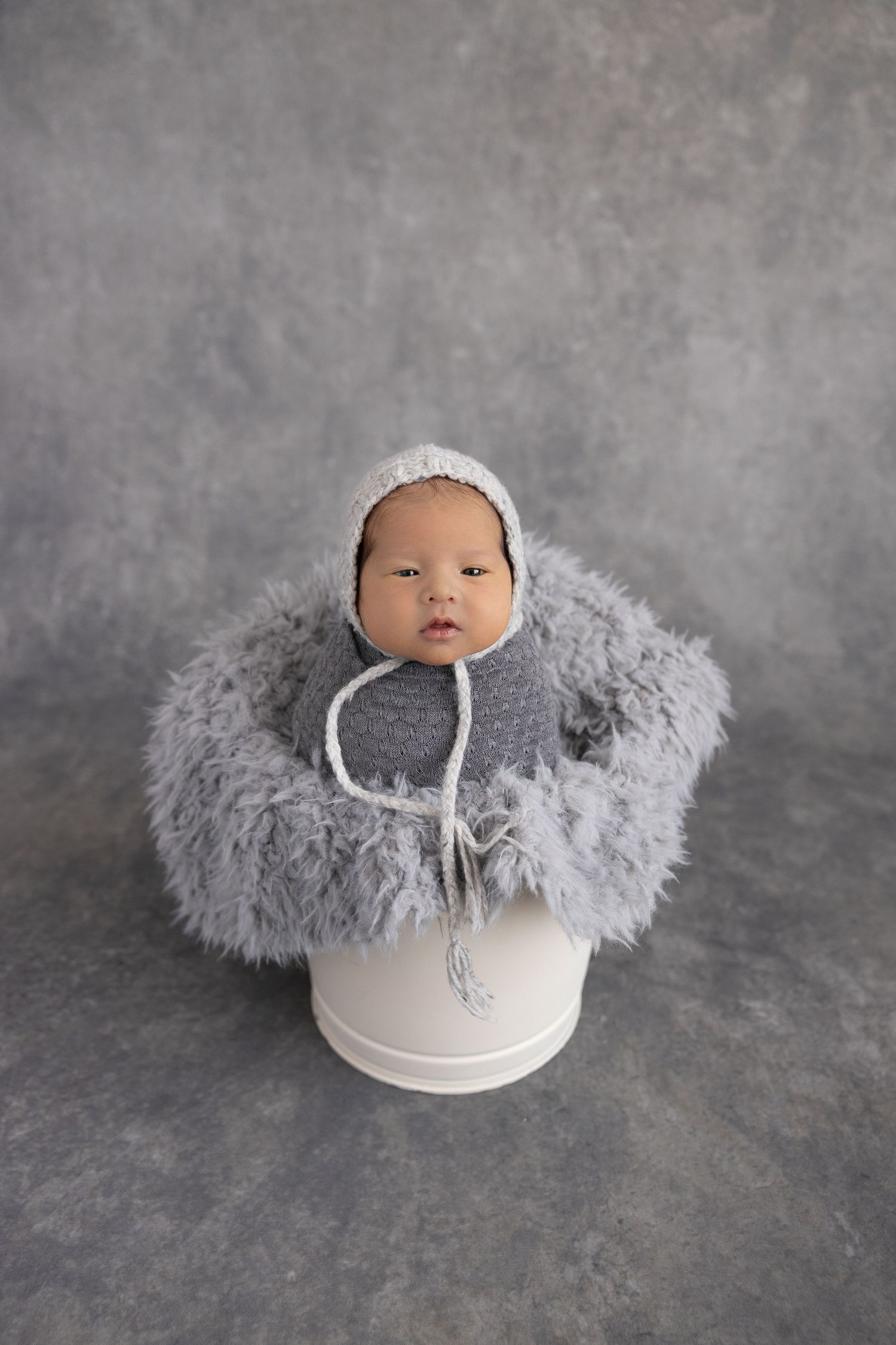 Tiny baby nestled in white bowl with soft grey fur accents on textured background.