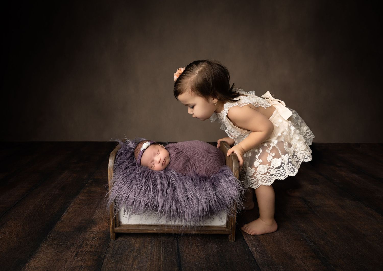 Two siblings interacting with purple cushion on wooden floor in studio setting.