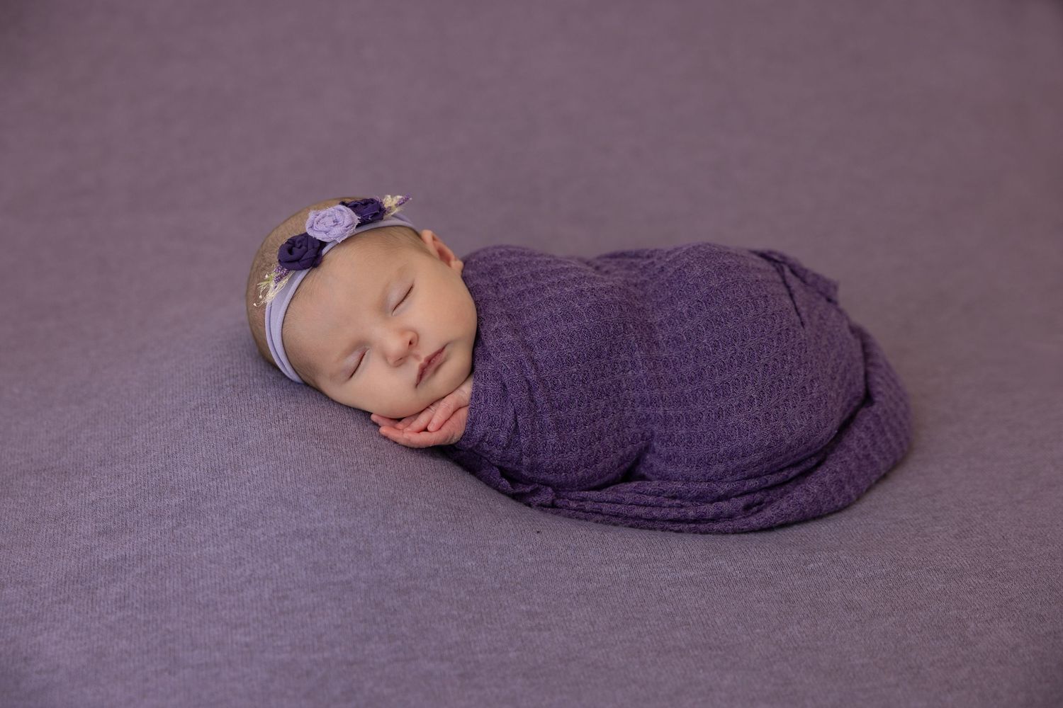 Sleeping newborn wrapped in purple fabric on textured violet background.