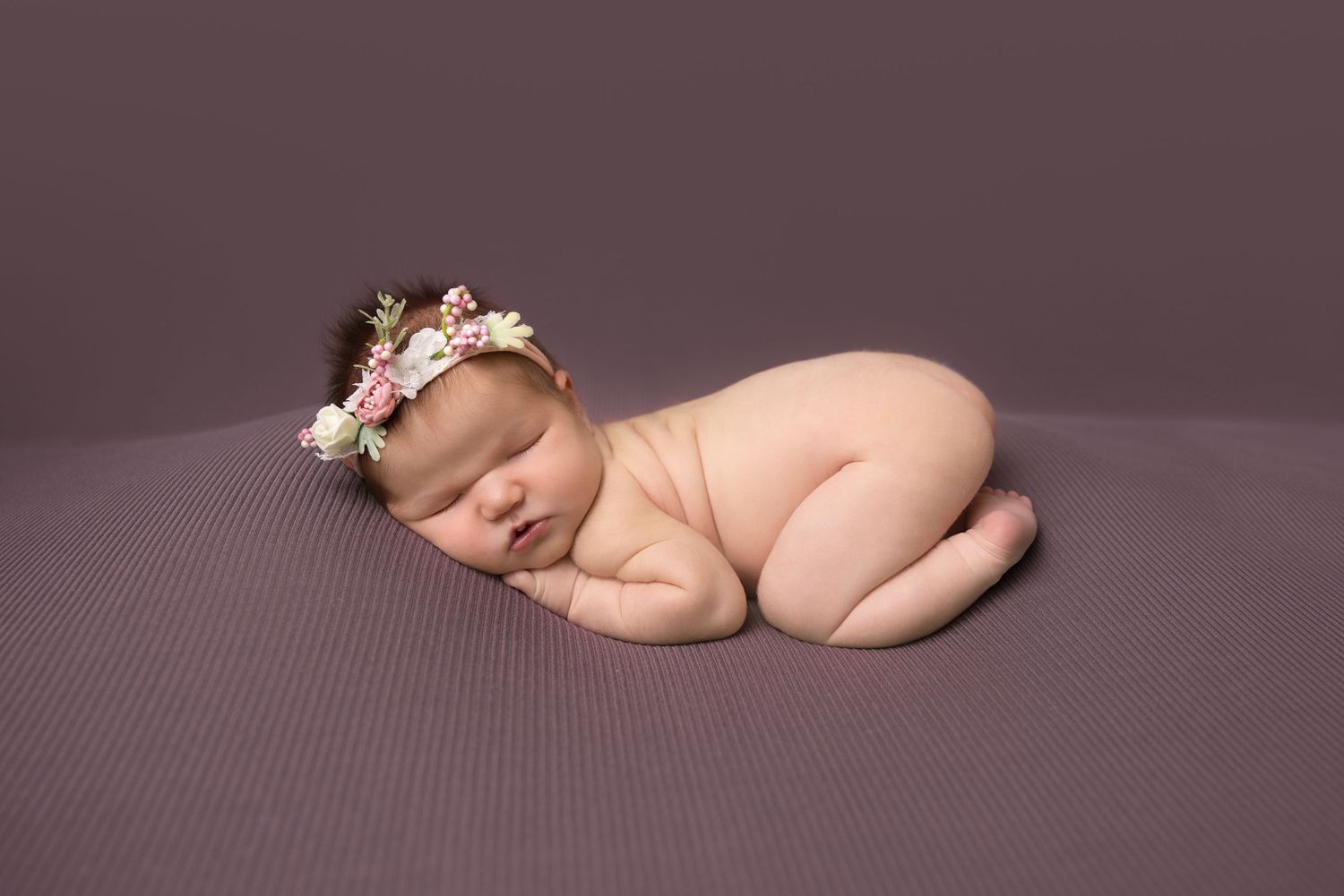 Newborn baby sleeping peacefully on plum colored blanket with delicate floral headband.