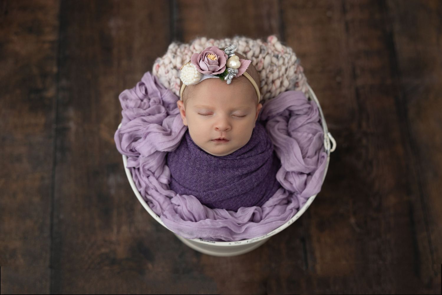 Newborn sleeping in purple wrap with floral headband in white bowl against dark wood background.