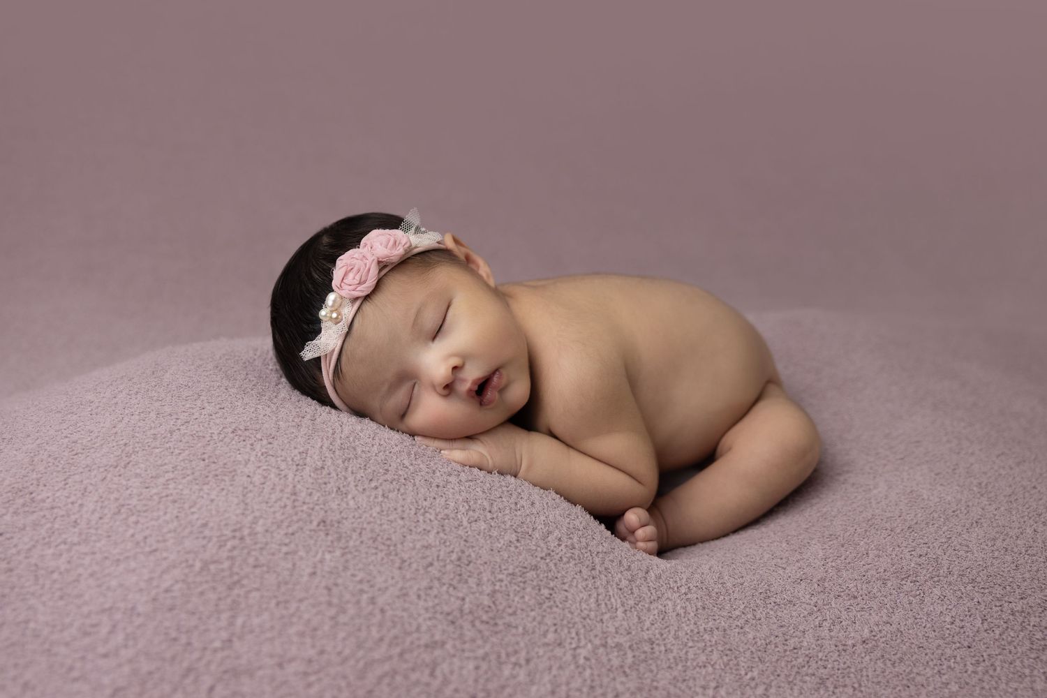 Peaceful sleeping newborn posed on soft mauve backdrop with delicate headband.