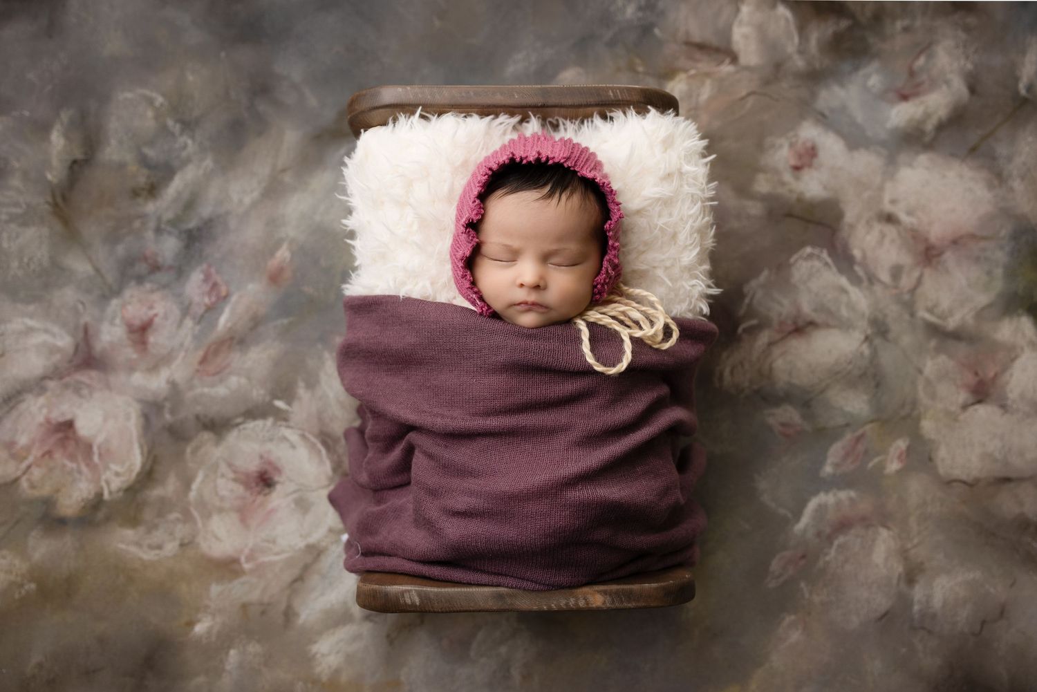 Infant sleeping in plum colored wrap on white fur with floral backdrop.