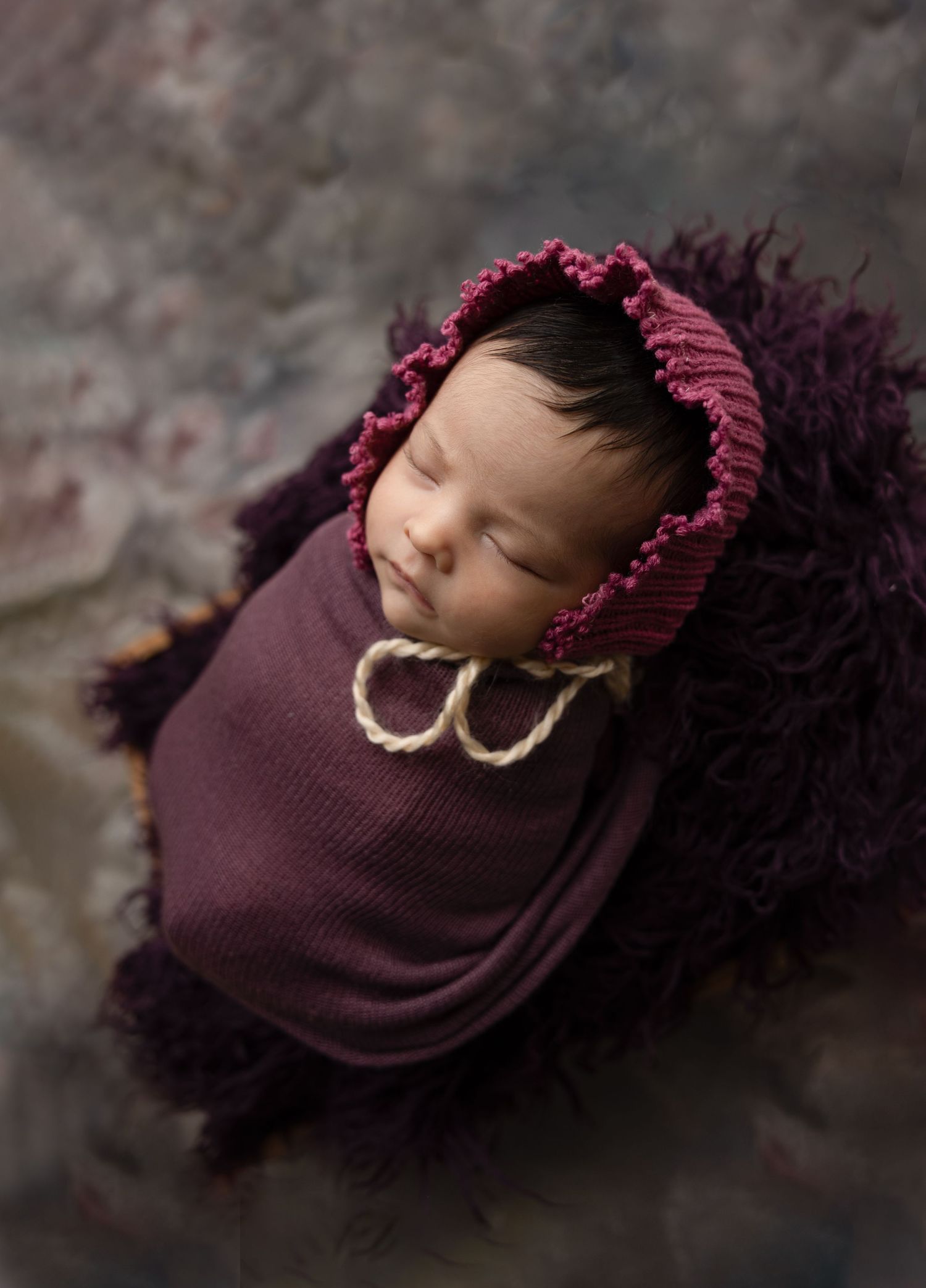 Newborn baby wrapped in burgundy fabric with matching textured bonnet.