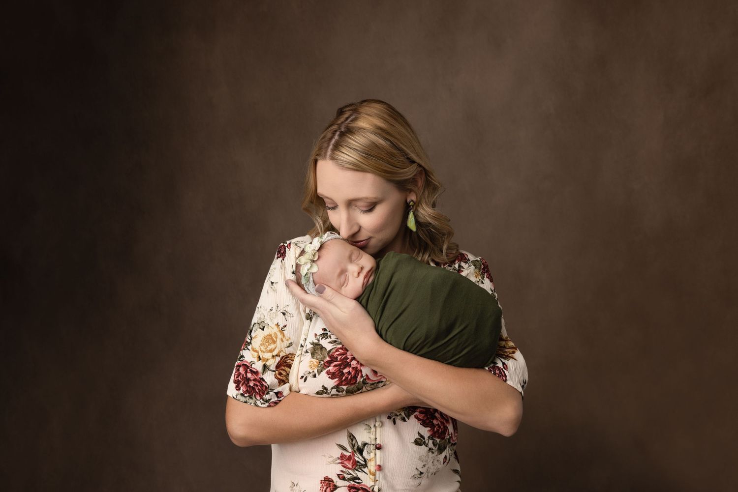 Tender portrait of woman in floral dress holding baby against dark brown background.