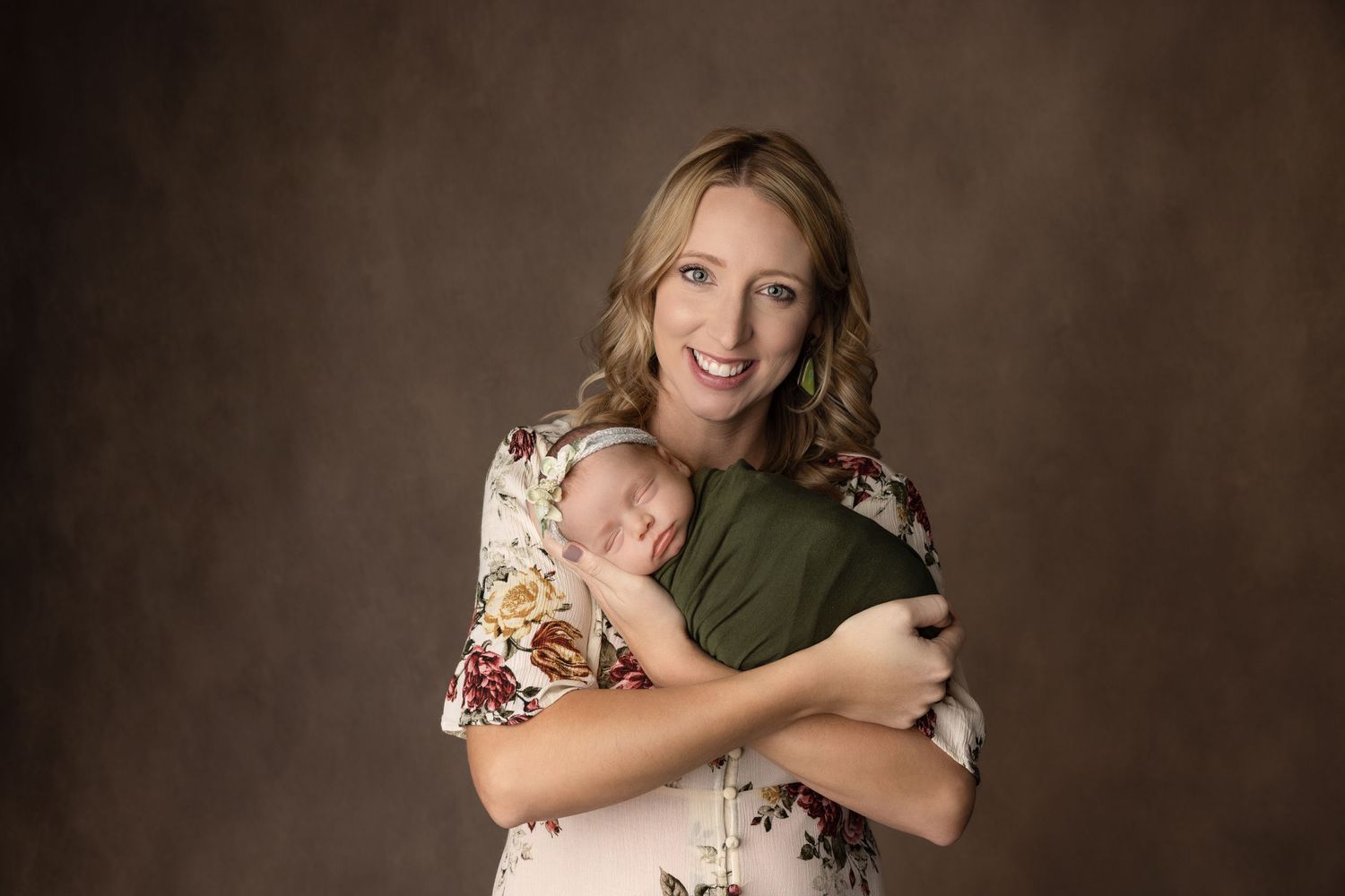 Mother in floral dress holding sleeping baby against dark brown studio backdrop.