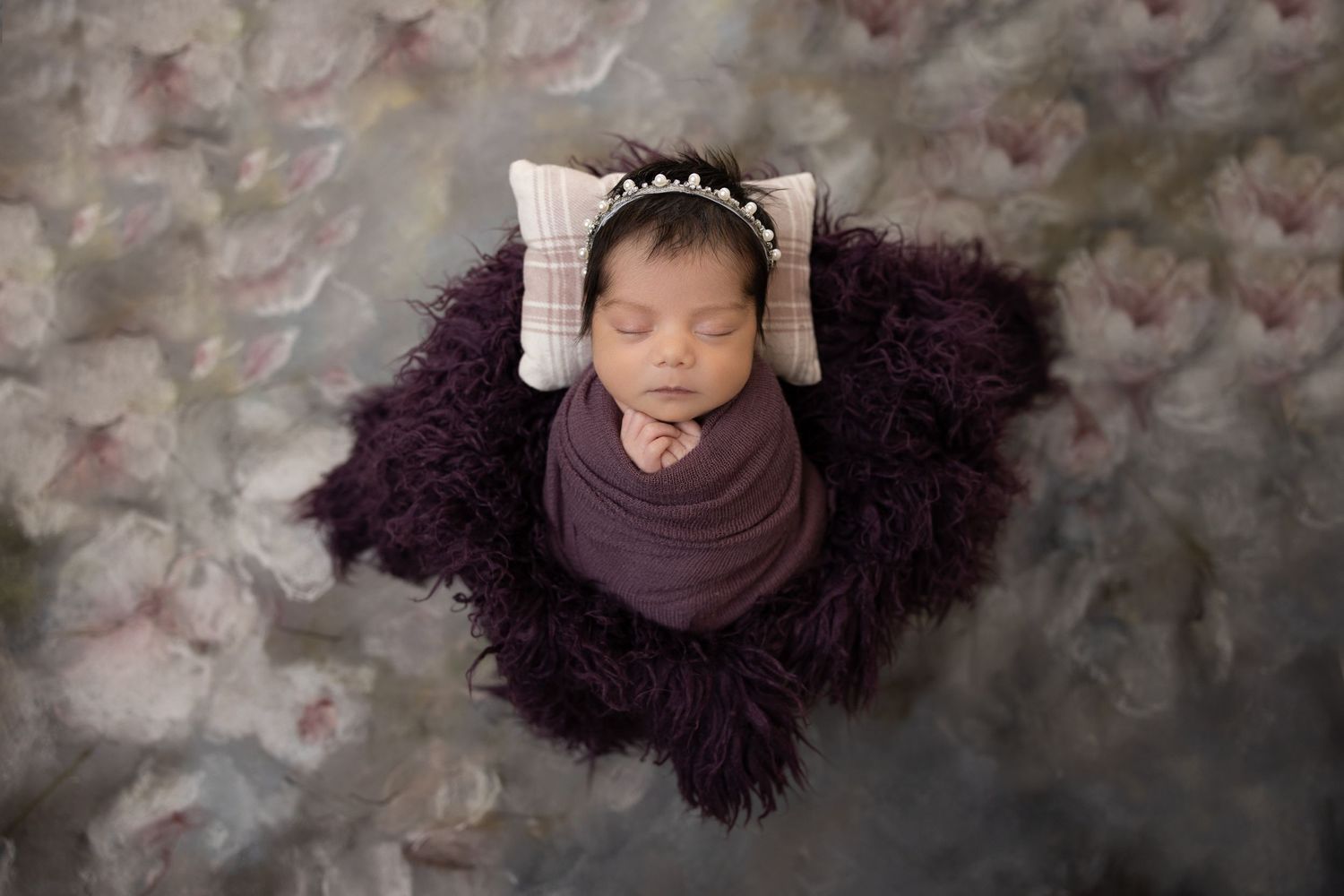 Sleeping newborn wrapped in purple fabric on dark fur with decorative white bow headpiece.