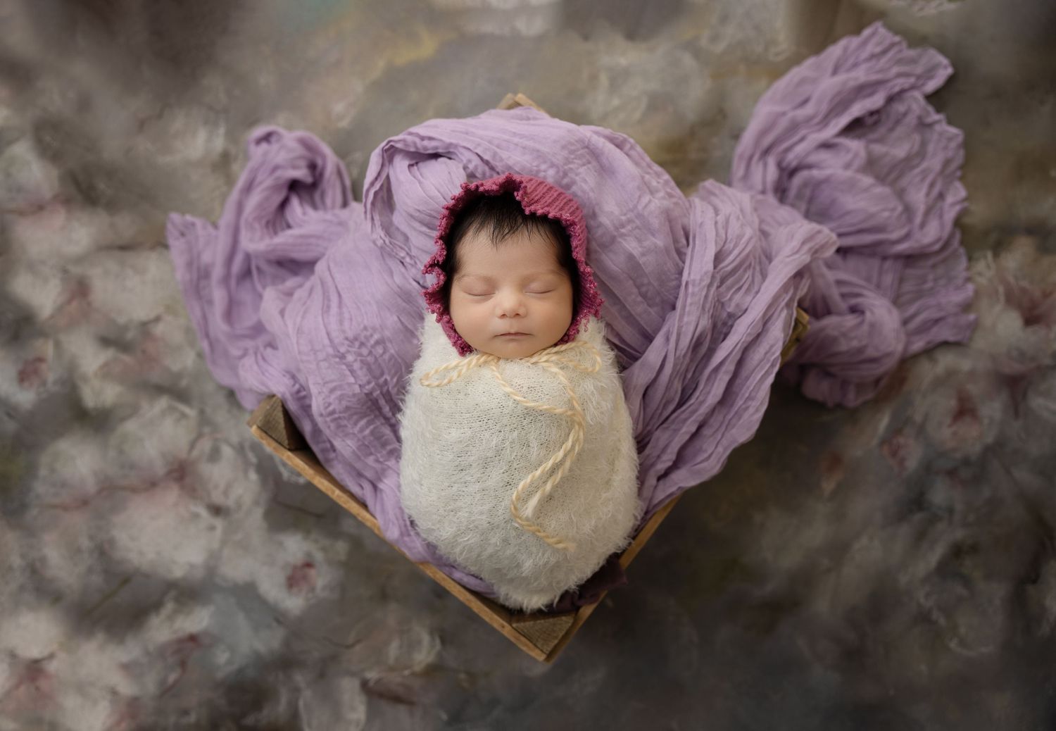 Sleeping newborn wrapped in white and surrounded by flowing purple fabric.