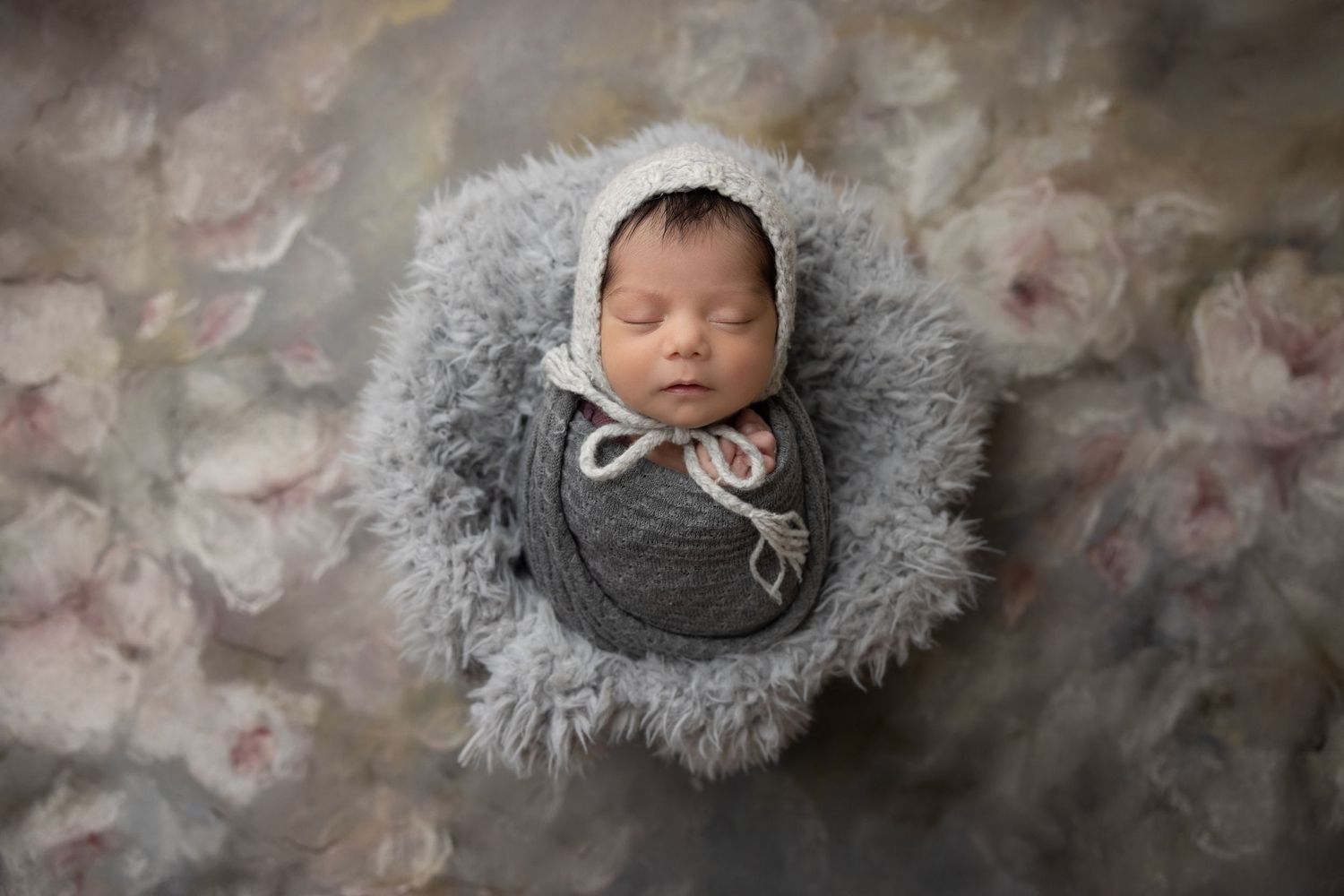 Sleeping infant wrapped in grey swaddle resting on fluffy grey fur against mottled background.