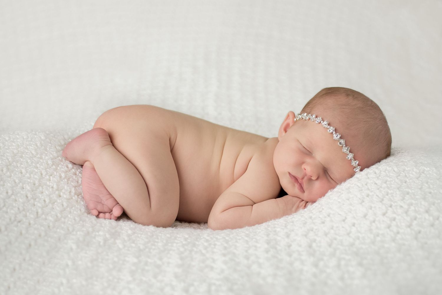 Sleeping newborn on textured white blanket with delicate pearl headband.