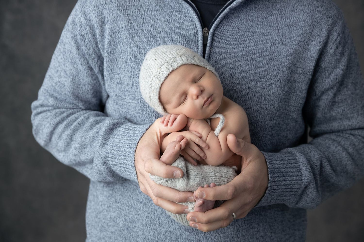Newborn baby in gray knitted wrap held gently in adult's arms during professional photography shoot.