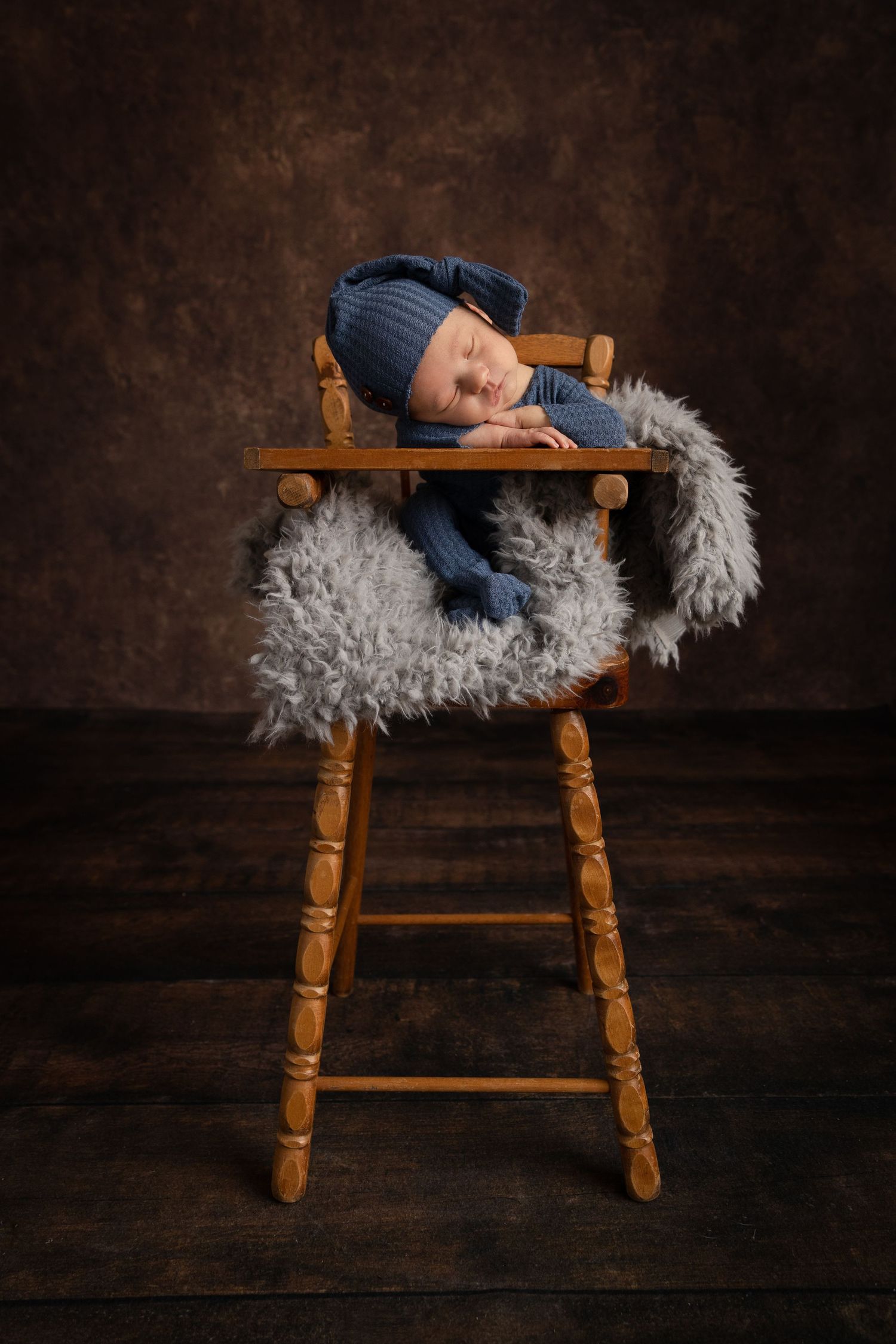 Sleeping baby in blue hat resting on wooden chair with fluffy gray cushion.