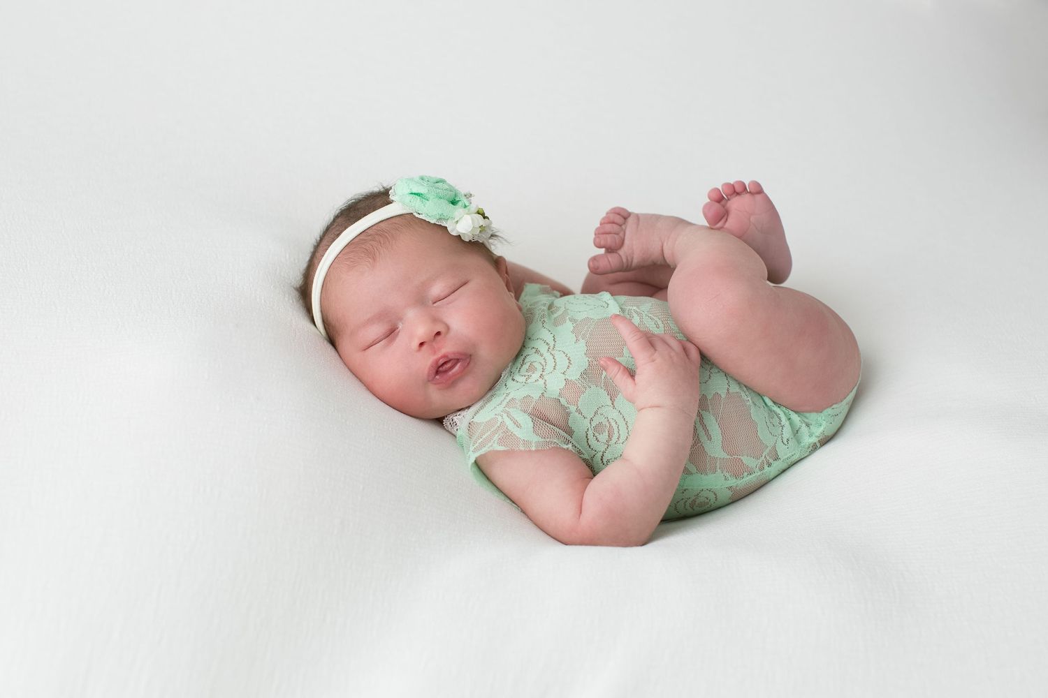 Newborn in mint green outfit with matching headband lying on white backdrop.