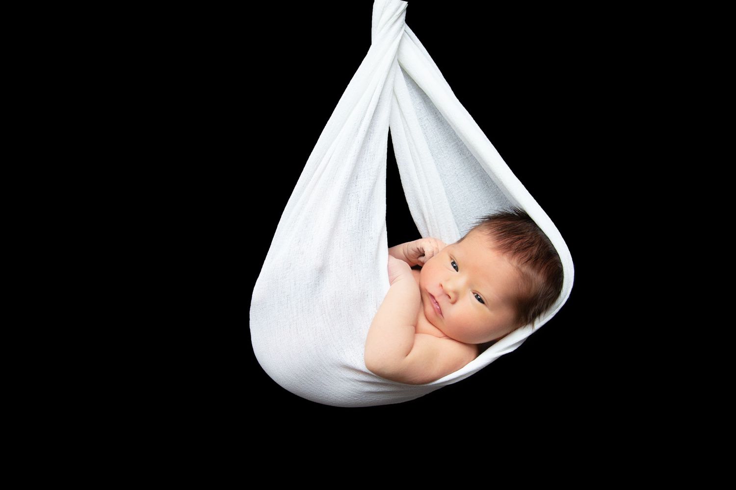 Minimalist newborn portrait with baby sleeping in suspended white fabric against black background.