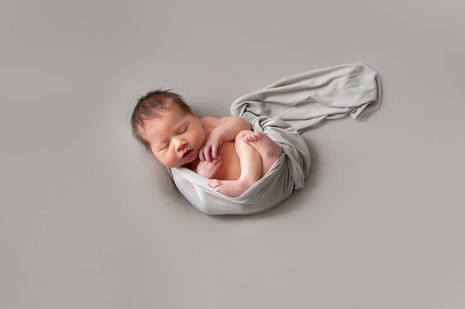 Newborn baby wrapped in soft gray fabric lying on pale backdrop during professional photo session.
