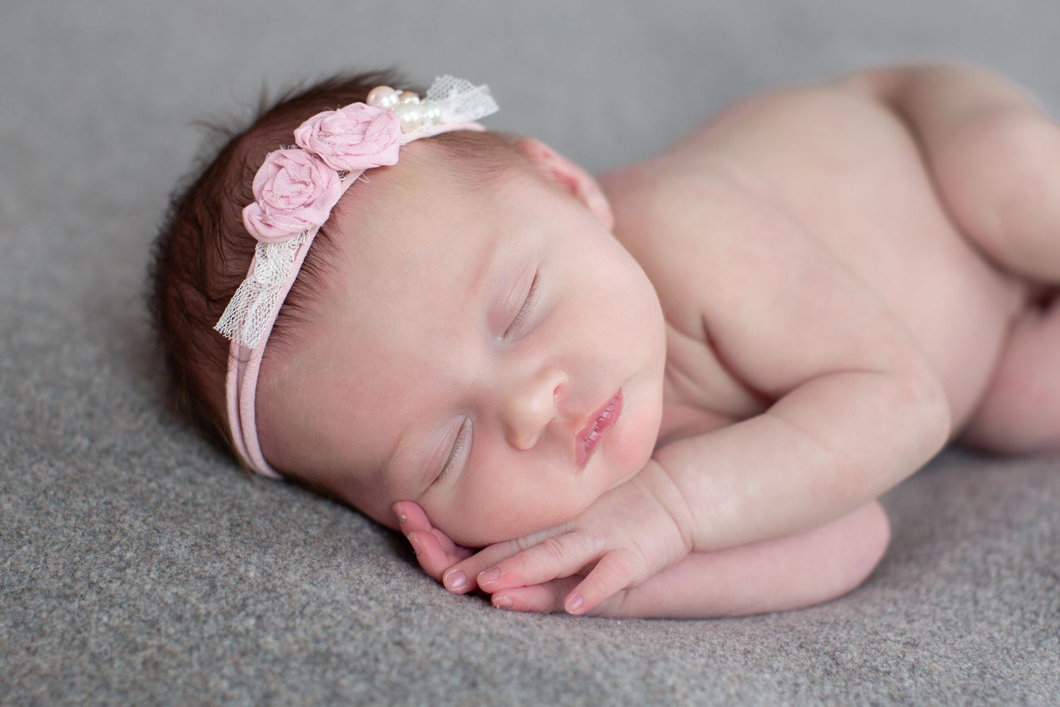 Sleeping newborn wearing pink headband resting peacefully on gray blanket.
