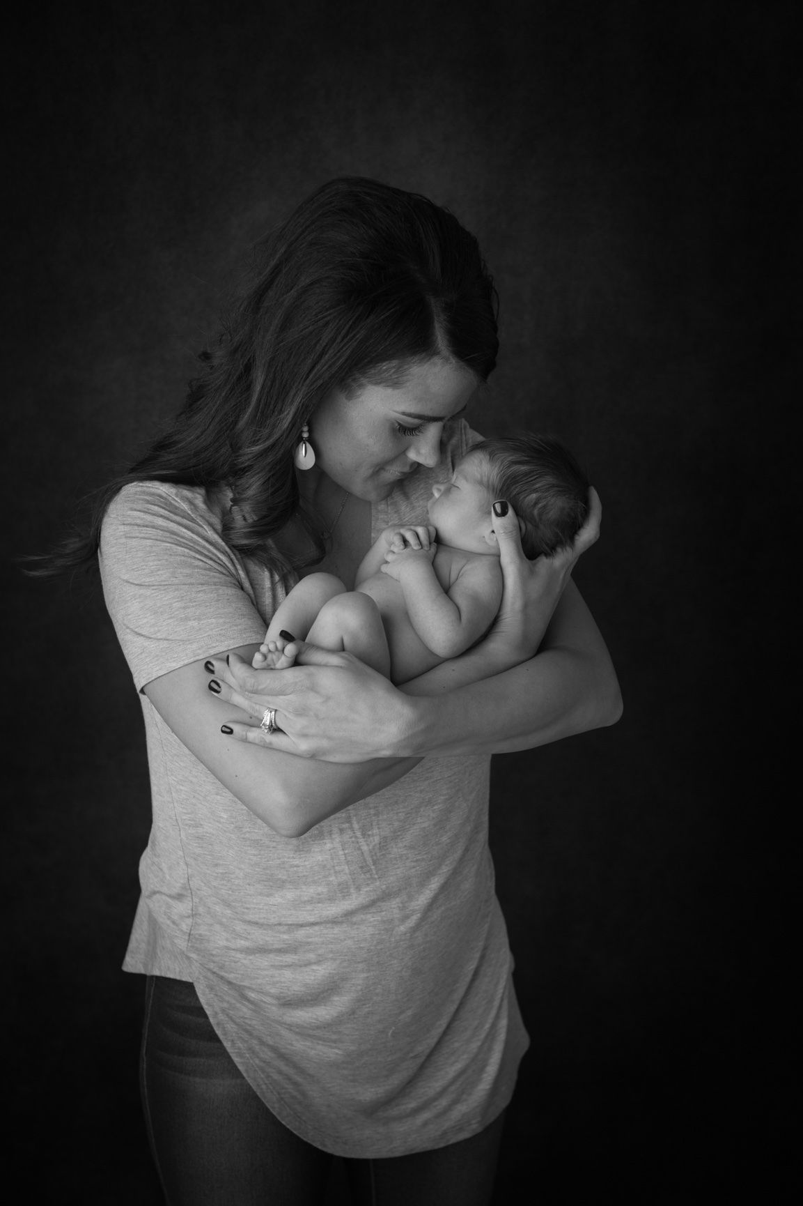 A tender black and white portrait of parent and newborn sharing an intimate moment against dark background.
