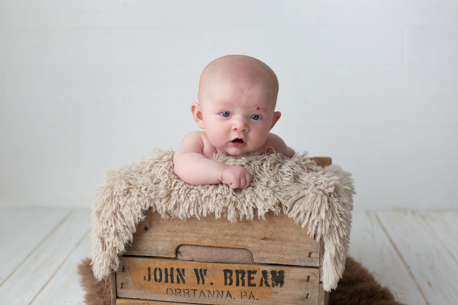 Baby resting on fluffy beige blanket in rustic wooden crate during studio session.