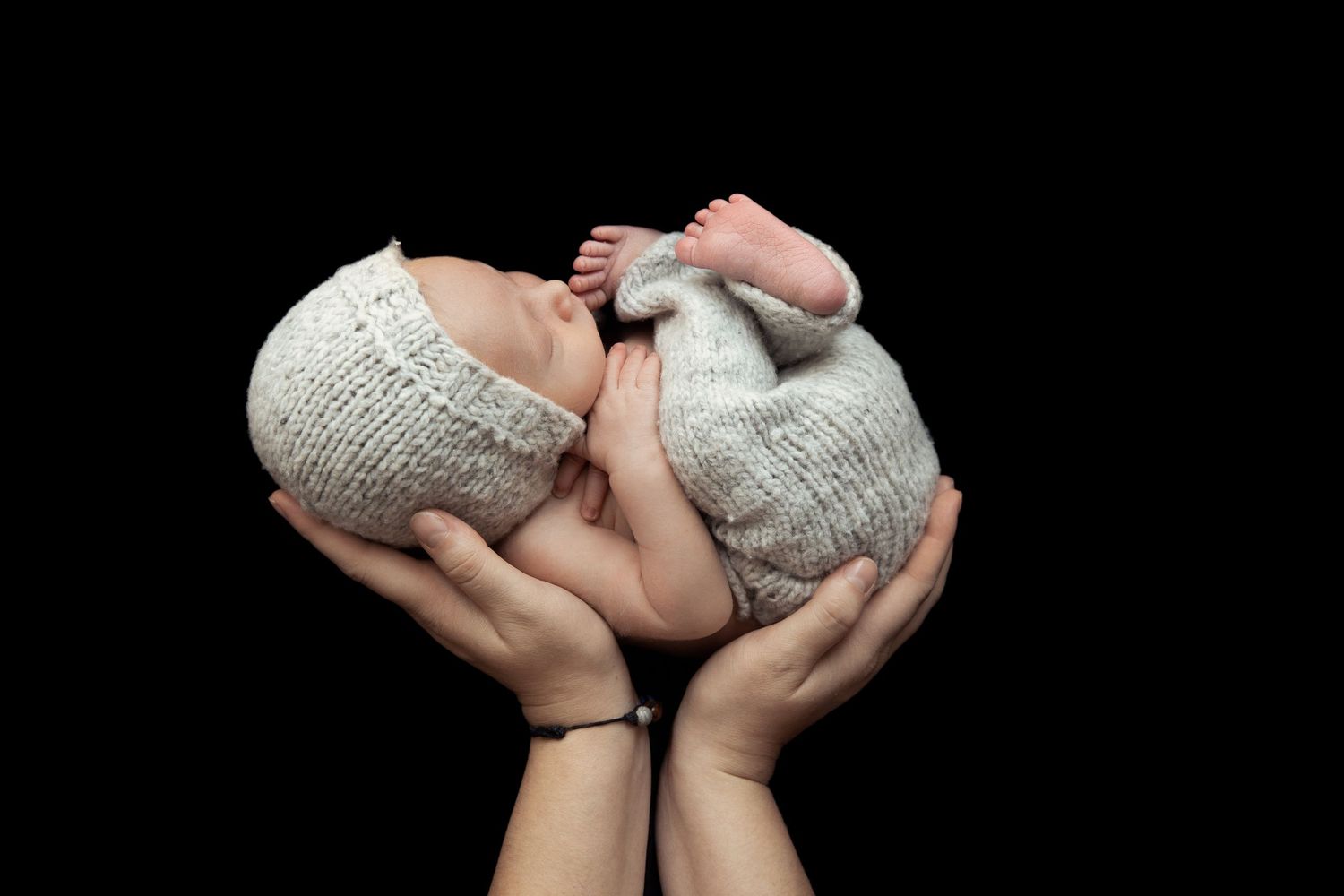 Hands gently holding newborn wrapped in gray knit blanket against black background.