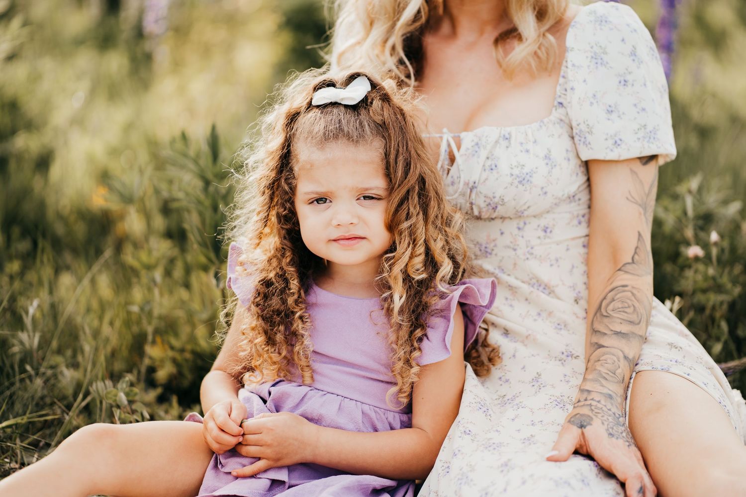 Young child with curly hair wearing a purple dress sits outdoors on a sunny day with her mom in a white dress.