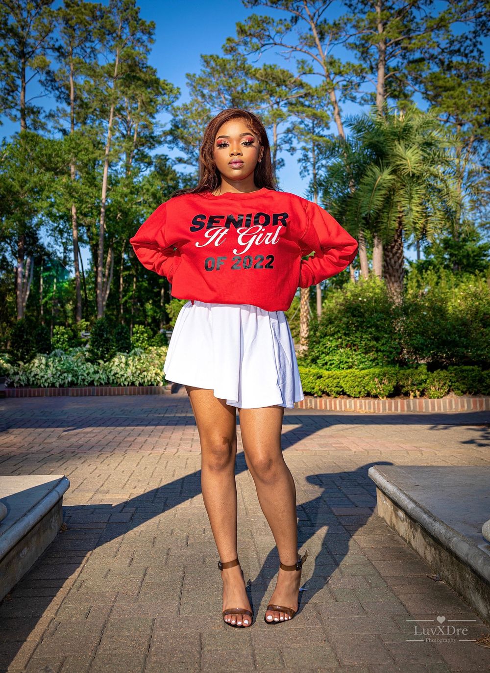 Senior wearing red sweatshirt and white skirt posing on brick pathway.