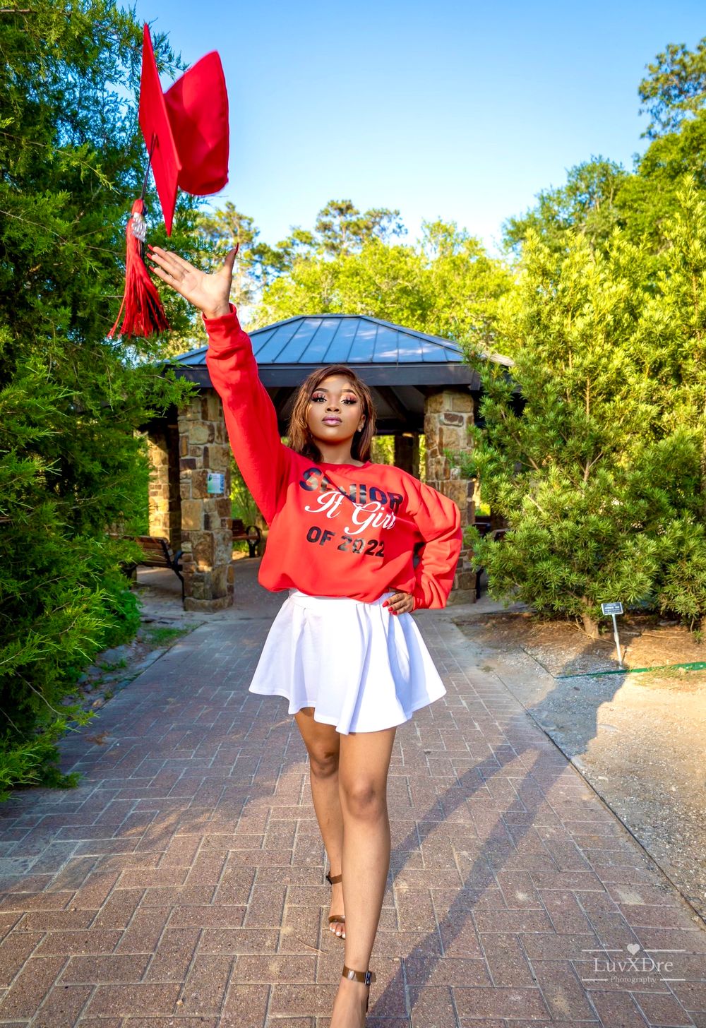 Graduate tossing red cap in celebratory pose on brick pathway with gazebo background.